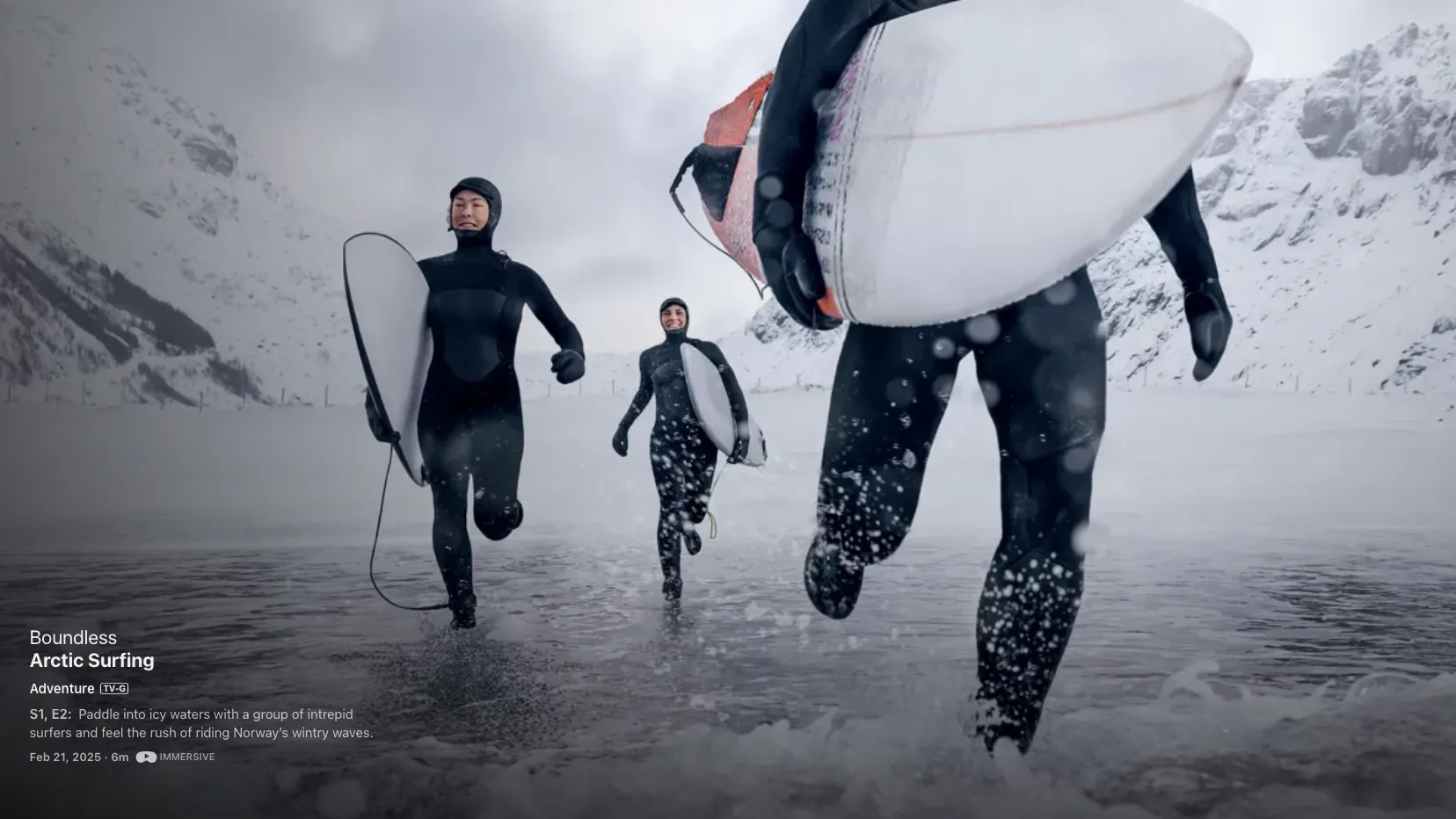 Three surfers in hooded wetsuits running into a grey Arctic sea below snow-covered peaks